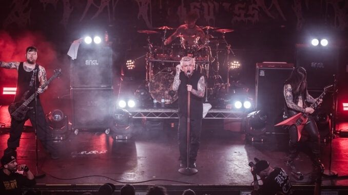 A metal band performing live on stage under bright spotlights with hazy smoke.